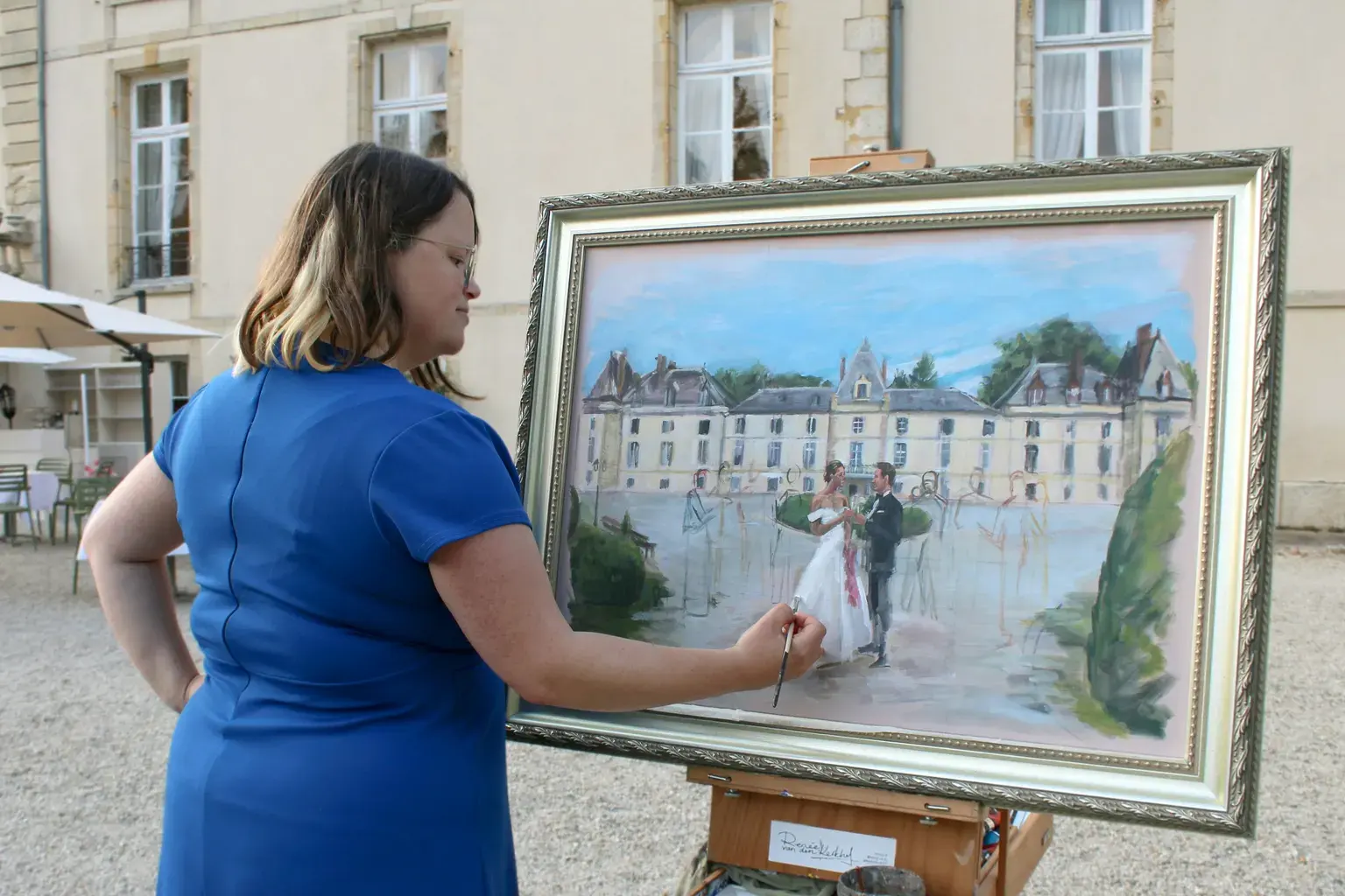 An artist in a blue dress paints a wedding couple on a large canvas outdoors.