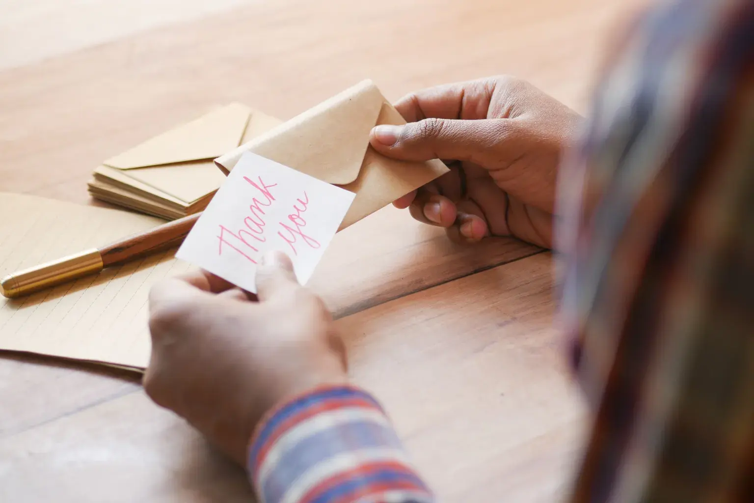 Photo by Towfiqu barbhuiya on Pexels Hands place a handwritten thank-you note into a kraft envelope on a wooden table.