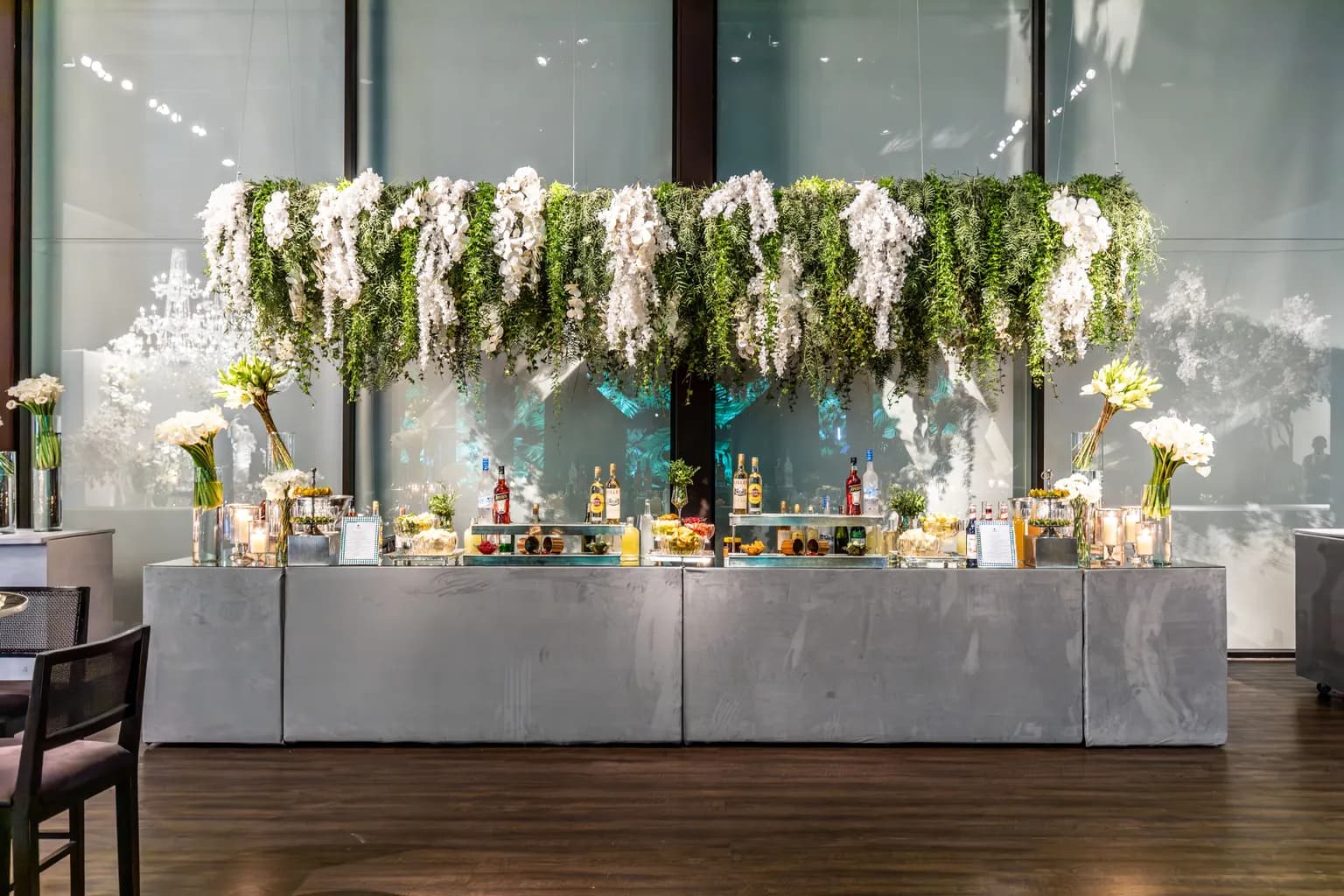Photo by Matheus Bertelli on Pexels A long gray event bar lined with liquor bottles, glassware and white orchids under a hanging canopy of greenery and white flowers.