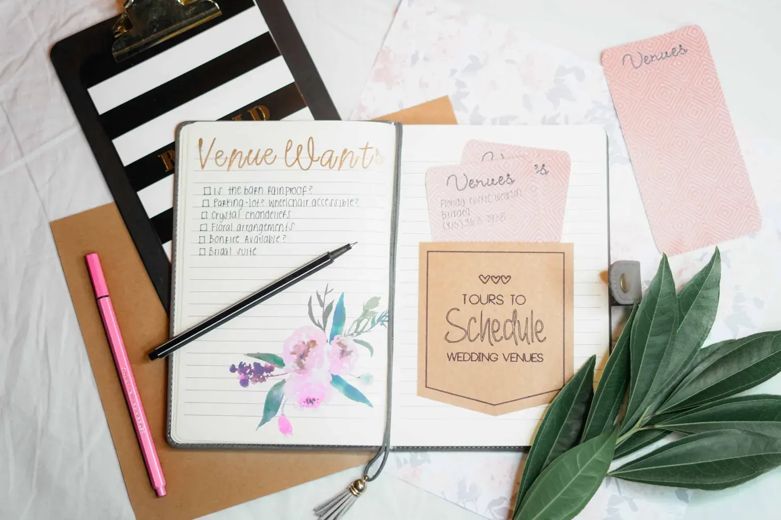 Photo by Natasha Fernandez on Pexels A wedding planning notebook with a venue checklist lies on a desk beside pens, cards, and green leaves.