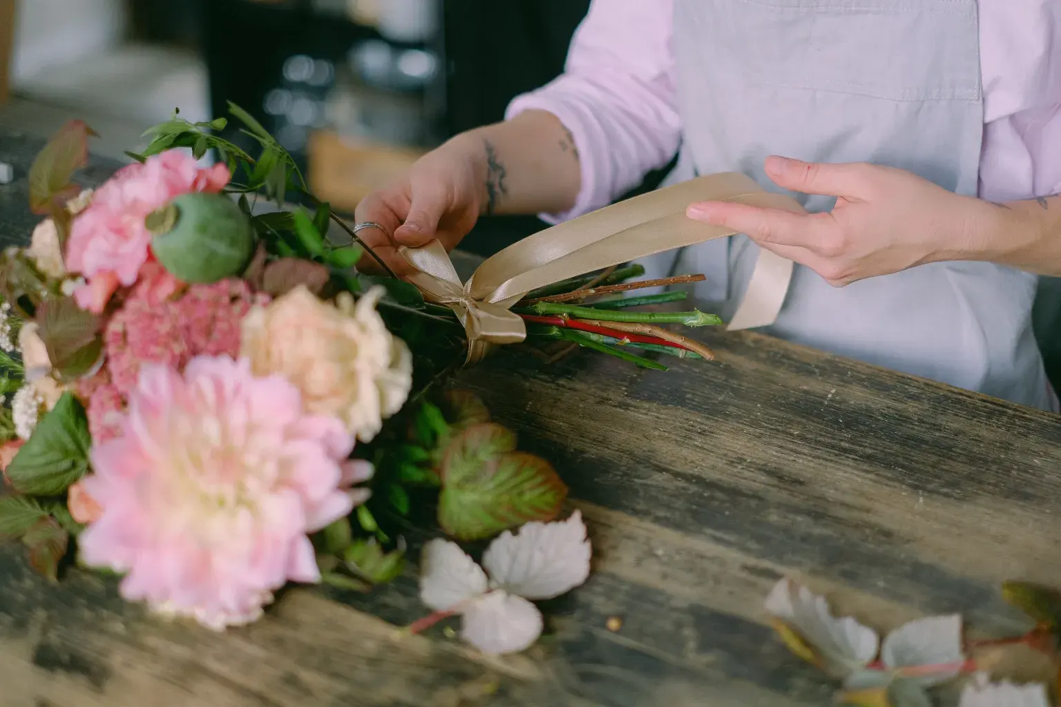 Photo by Anna Shvets on Pexels A florist ties ribbon around a bouquet of pink and cream flowers on a wooden work table.