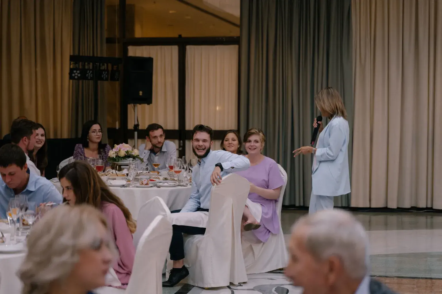 Photo by Agafonova Photo on Pexels Guests sit at round reception tables in a ballroom while a speaker holds a microphone.