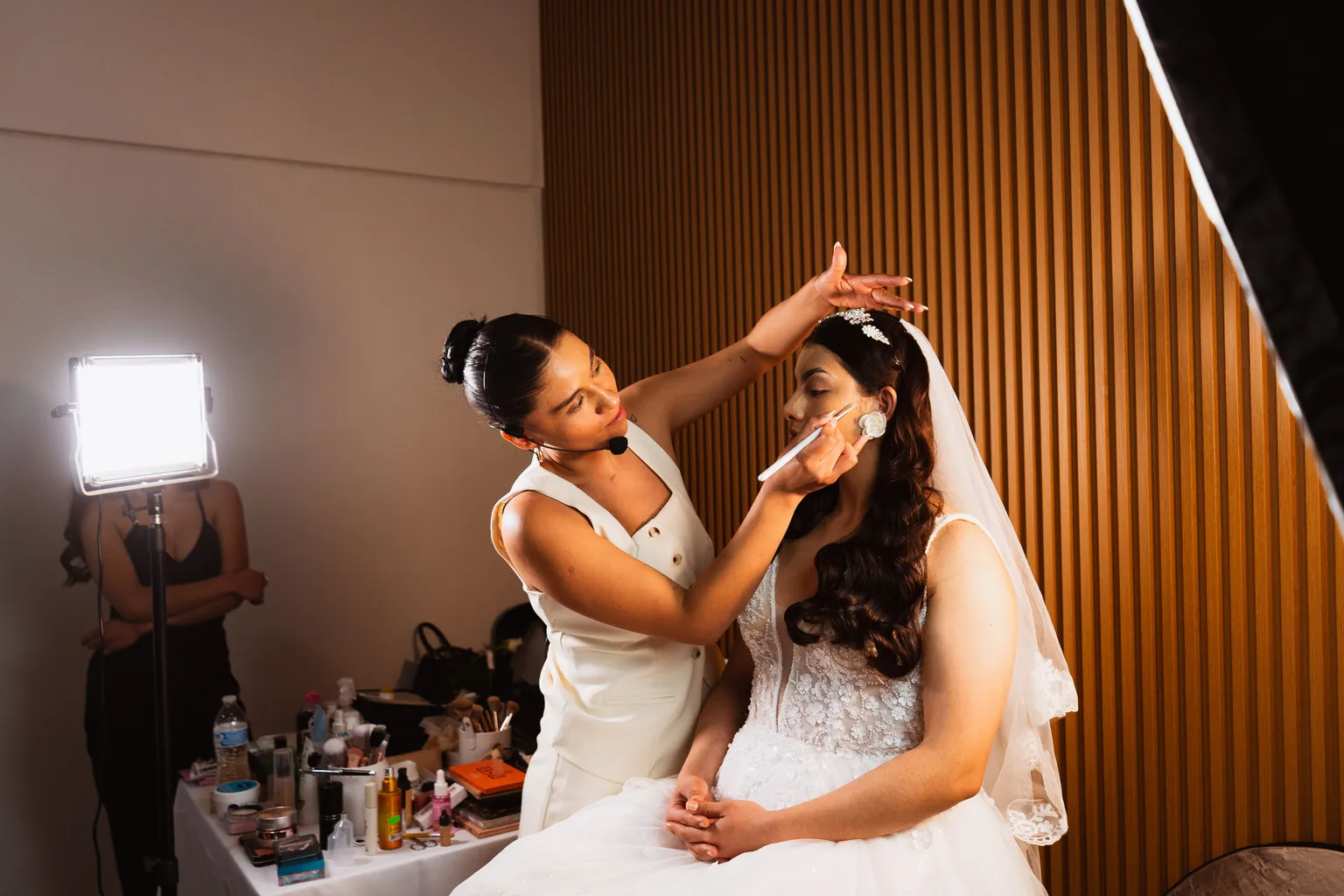 Photo by Vladimir Flores on Pexels A makeup artist in a white vest brushes makeup onto the cheek of a seated bride in a lace gown, veil, and tiara, beside a table of cosmetics and a studio light.