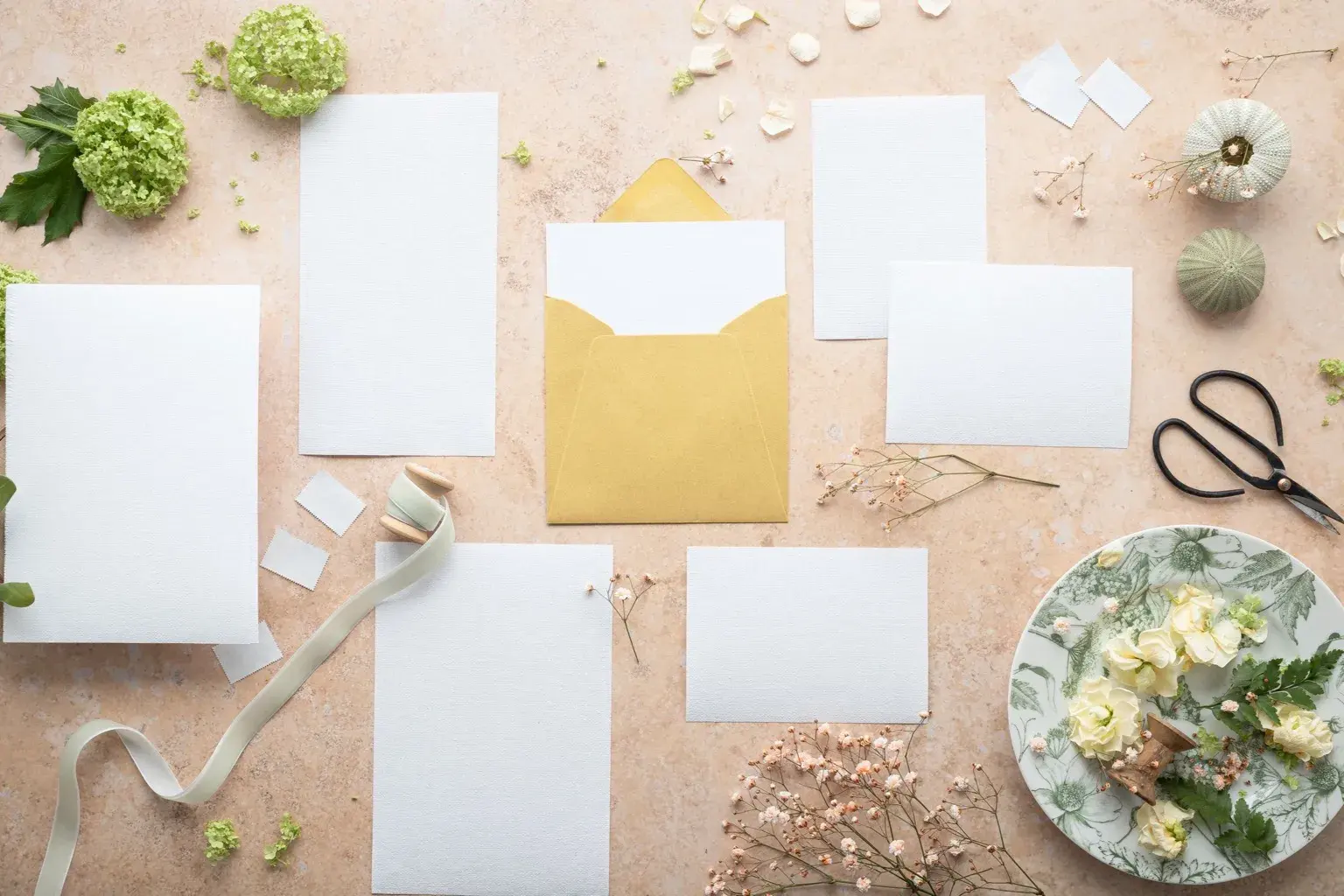 Photo by Micheile Henderson on Pexels Blank invitation cards and a gold envelope are arranged with flowers, ribbon, and scissors on a peach work surface.