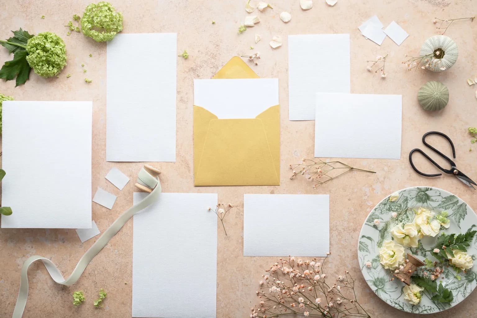 Photo by Micheile Henderson on Pexels Blank invitation cards and a gold envelope are arranged with flowers, ribbon, and scissors on a peach work surface.