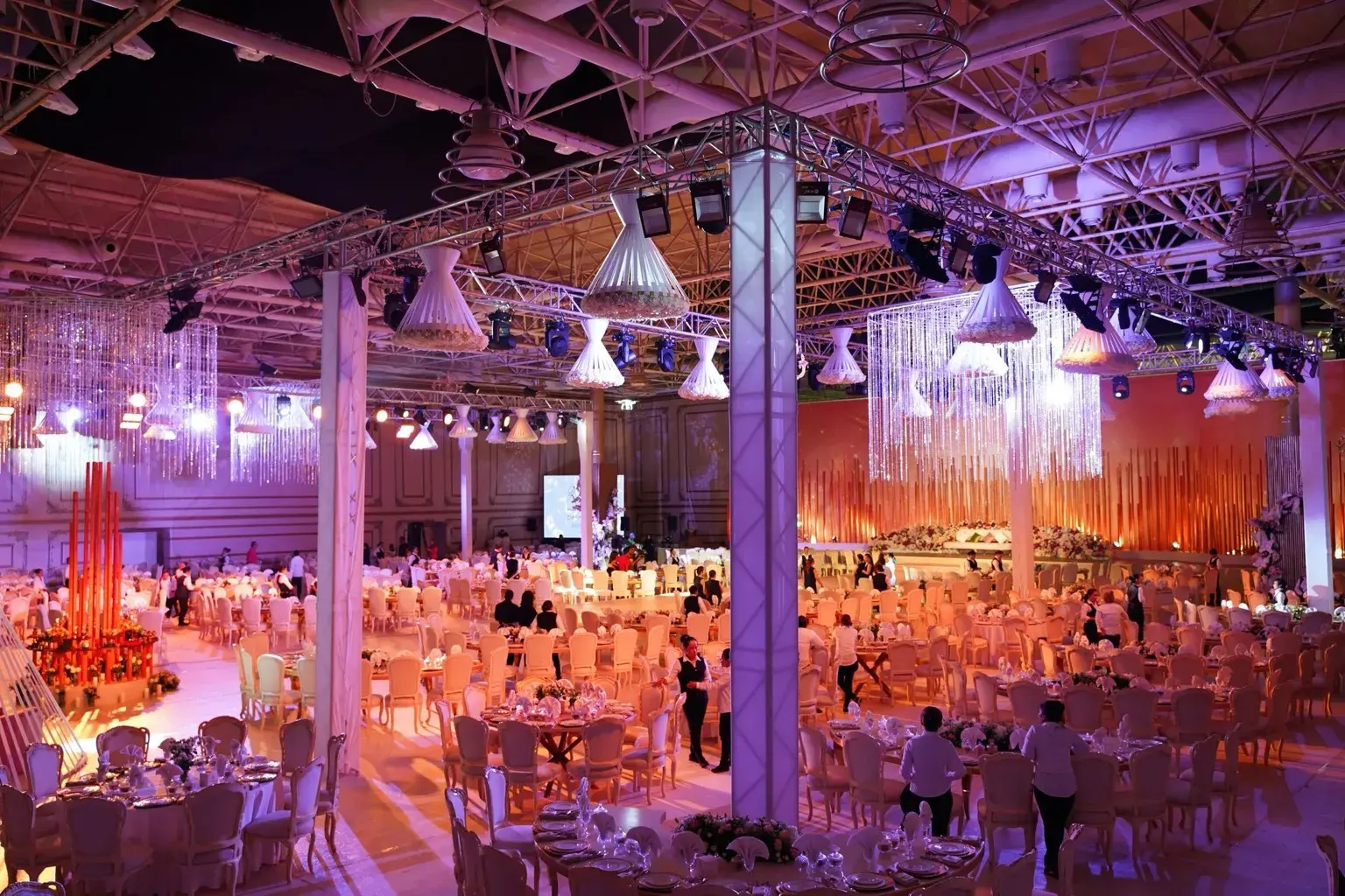 Photo by Denys Gromov on Pexels A large reception hall with tables is lit by purple wall lights and hanging fixtures.
