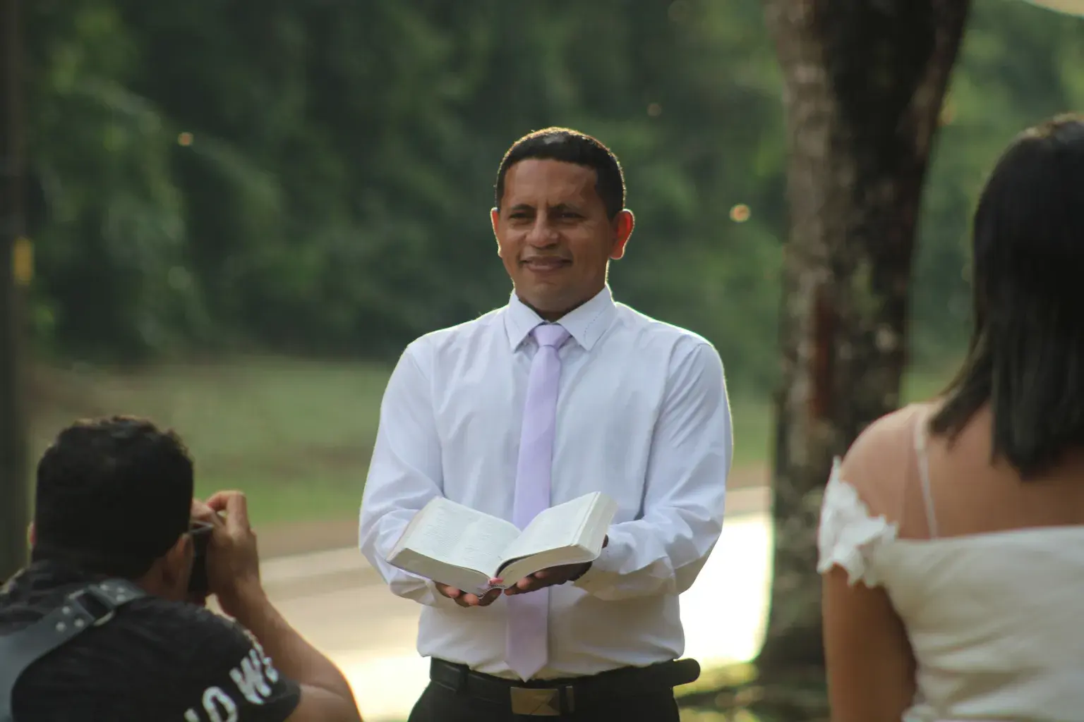 Photo by Gustavo Santana on Pexels An officiant in a white shirt and tie holds an open book during an outdoor ceremony while a photographer works nearby.