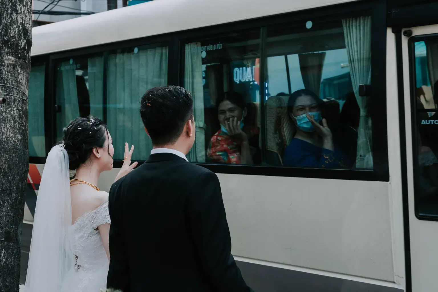 Photo by HONG SON on Pexels A bride waves to passengers through a bus window while a groom stands beside her.