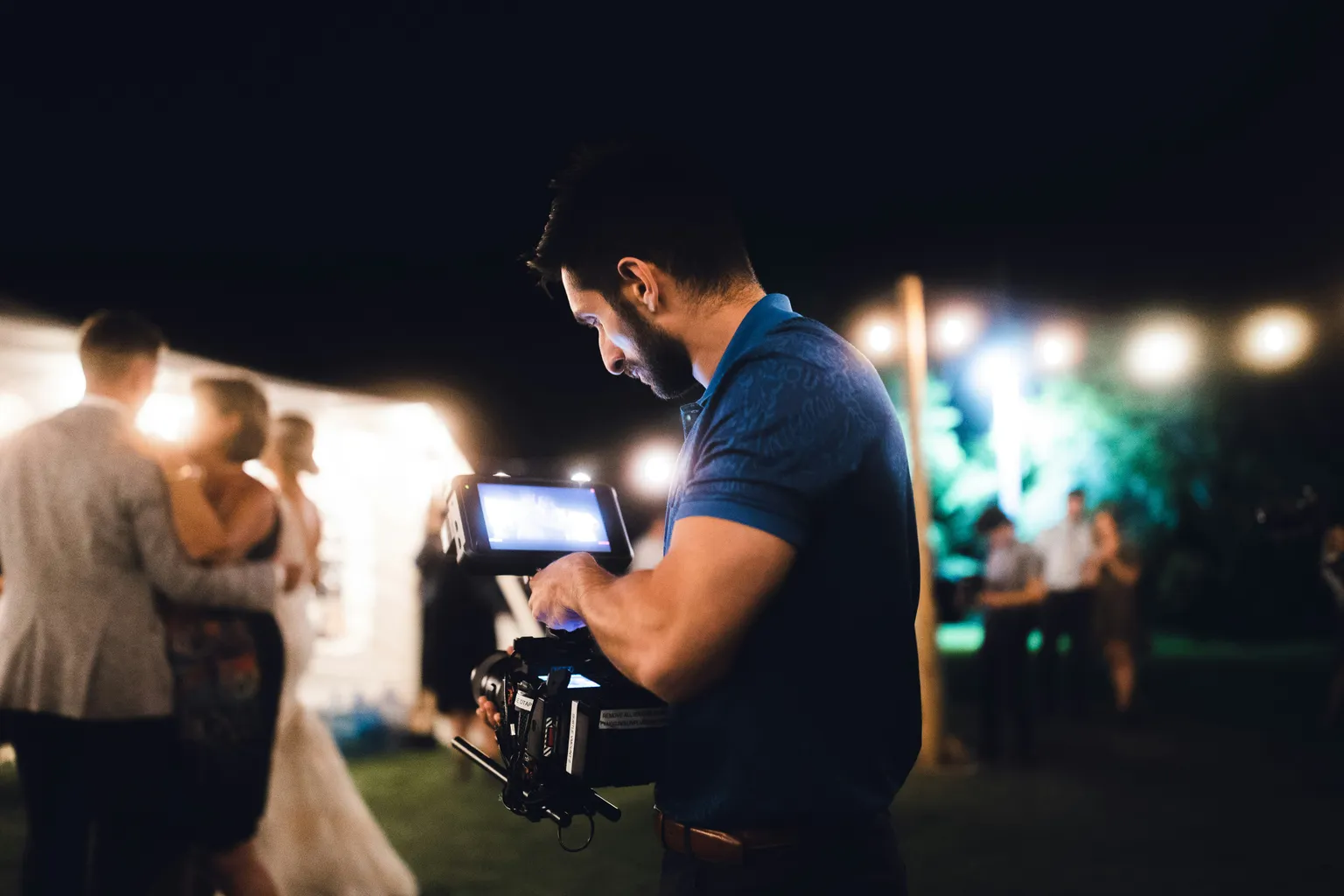 Photo by Anastase Maragos on Unsplash A videographer in a blue polo checks the monitor on his handheld cinema camera at a nighttime outdoor reception, with a couple dancing and string lights blurred behind him.